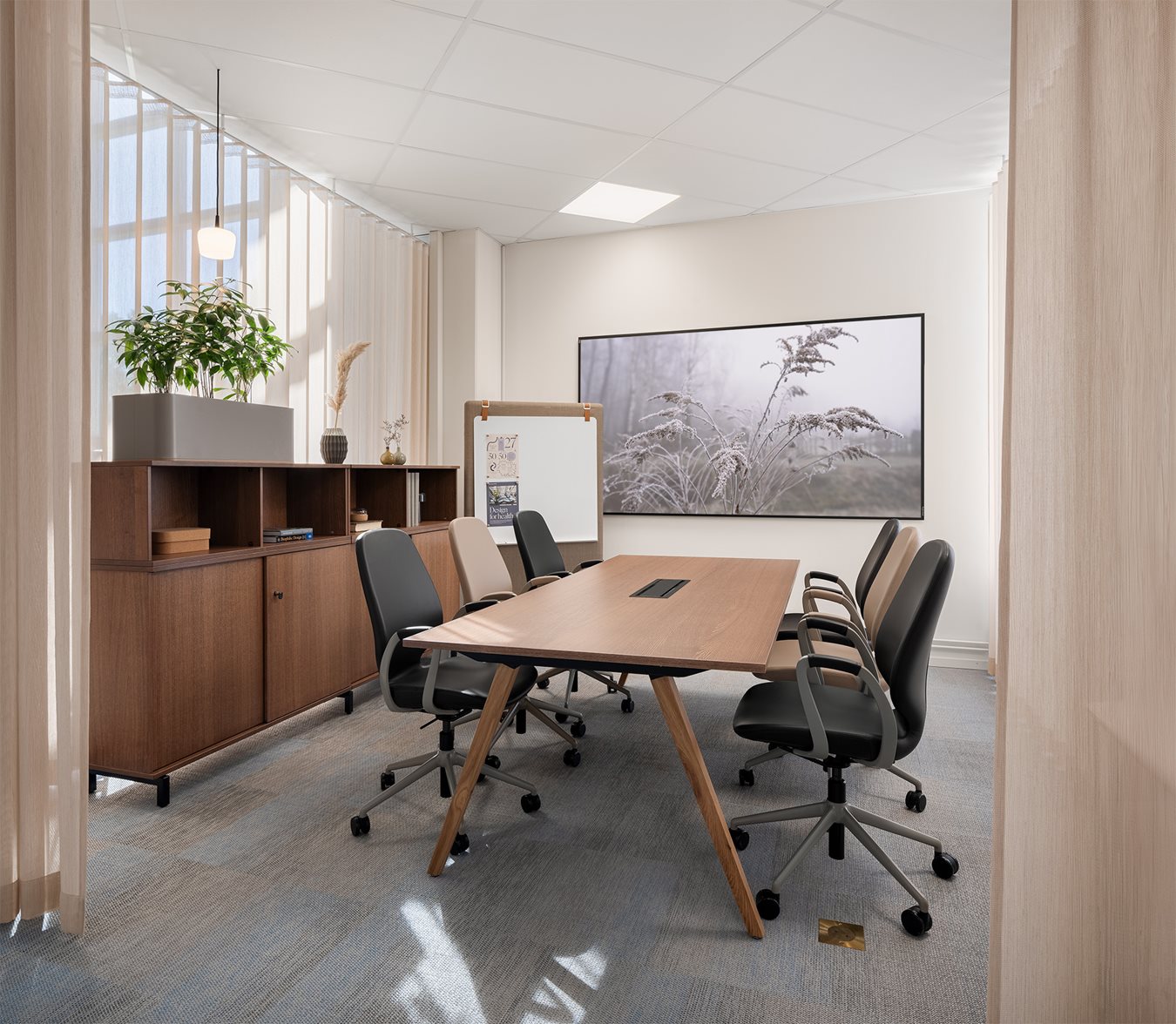 A modern conference room with a rectangular wooden meeting table, black and beige office chairs with wheels, a large conference room screen on the wall, and a storage unit with plants. The room has light-colored walls, a textile carpet, and soft lighting.