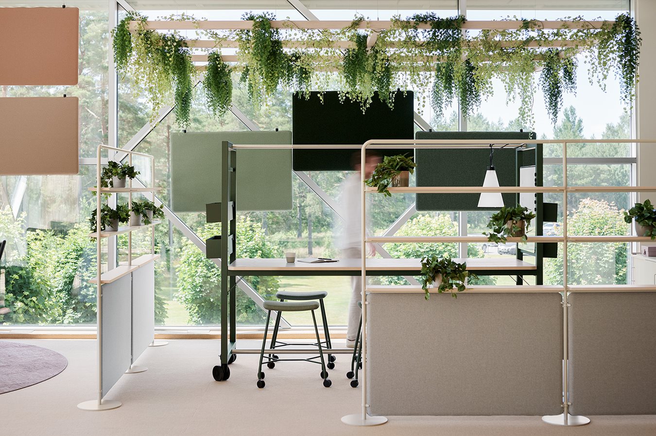 An open and flexible project space with a large meeting table, bright ceiling suspended screens, floor screens, and lush greenery hanging from the ceiling. Large windows let in natural light and provide a view of the greenery outside.